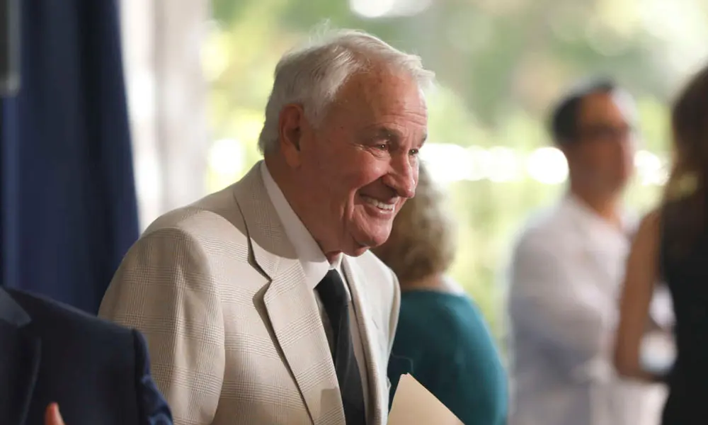 Tom Golisano smiles while holding a folder as people mingle around him.