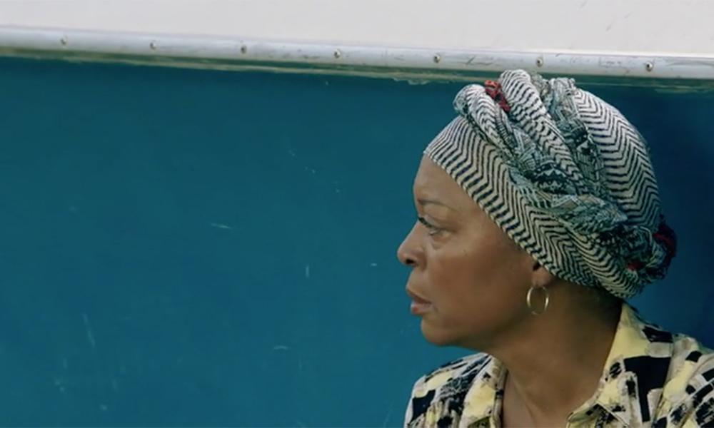 Screenshot of Black actress Tina Lifford, wearing a colorful head wrap and standing in profile against a bright blue wall.